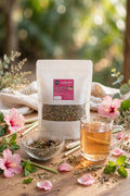 A stand-up pouch of herbal tea with a clear window showing loose dried herbs, styled on a wooden surface with a glass of brewed tea, a small bowl of loose herbs, and pink flowers in a soft outdoor setting.